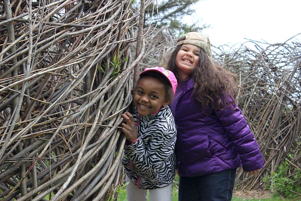 Two children in hats and jackets beside a woven wooden sculpture